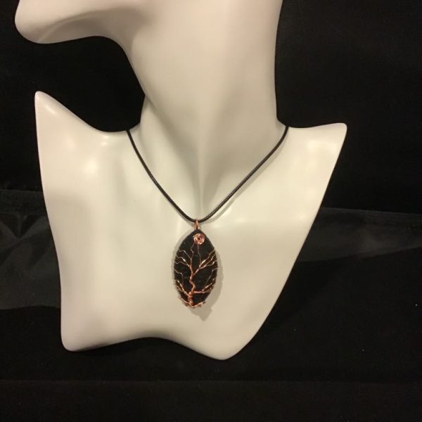 The Midnight Enchantment Necklace features a dark, oval pendant with intricate copper wirework in a tree of life design, hanging from a sleek black cord. Perfect for adding a touch of mystical elegance to any outfit.